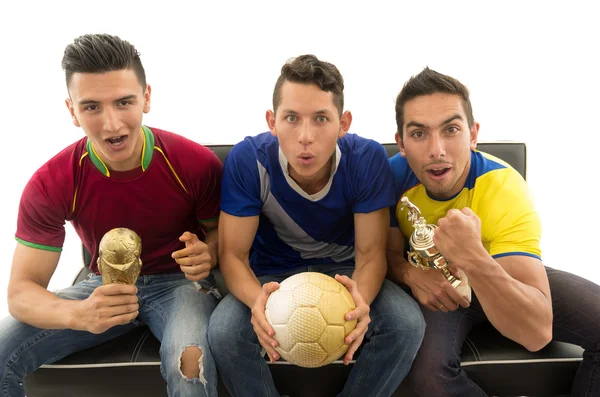 Three friends sitting on sofa wearing sports shirts, holding ball and trophy screaming cheering at camera with enthusiasm, white background, shot from above — Stock Photo, Image
