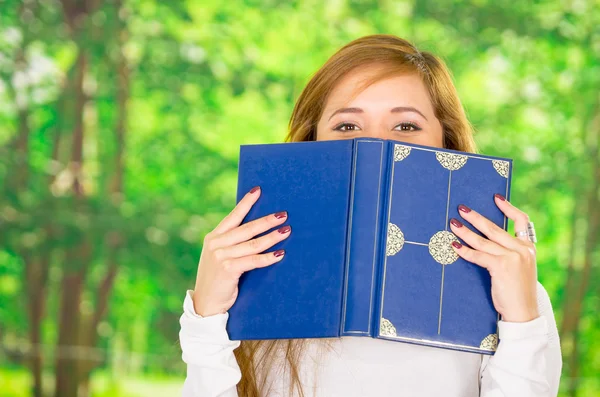 Headshot jonge brunette die betrekking hebben op de helft van het gezicht met blauwe boek, Tuin achtergrond — Stockfoto