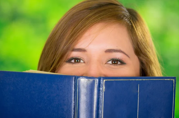 Kopfschuss junge Brünette bedeckt Hälfte des Gesichts mit blauem Buch, Garten Hintergrund — Stockfoto