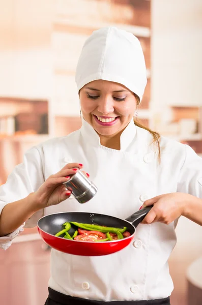 Mujer chef sosteniendo sartén roja con verduras picadas en el interior, añadiendo sal del recipiente de metal — Foto de Stock