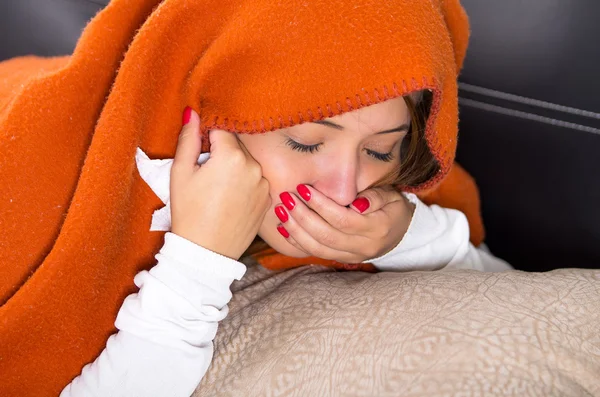 Morena acostada debajo de una manta naranja y cubriéndose la boca con la mano, enferma de gripe — Foto de Stock