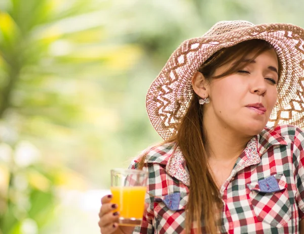 Mujer joven en camisa de patrón cuadrado disfrutando de un jugo amarillo, ambiente de jardín — Foto de Stock