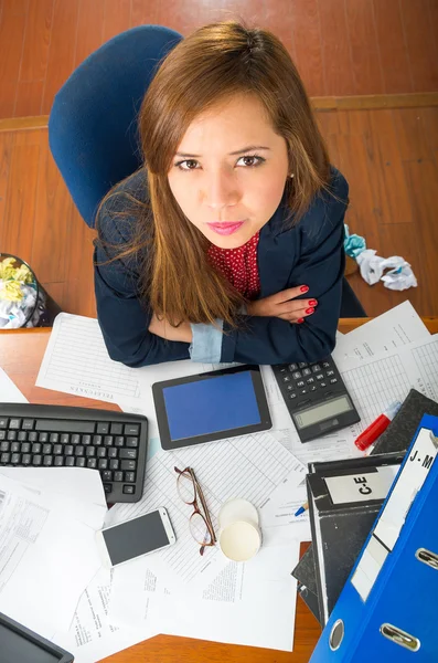 Ung kvinna sitter vid skrivbord med levererar utspridda, samverkande tittar in i kameran som sett från ovan vinkel — Stockfoto