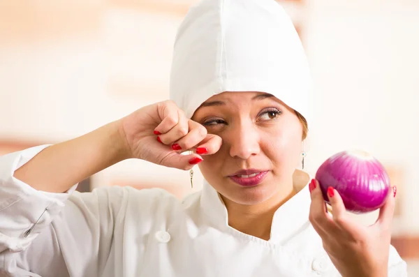 Mujer chef disparo en la cabeza sosteniendo una cebolla roja y limpiando las lágrimas con otra mano — Foto de Stock