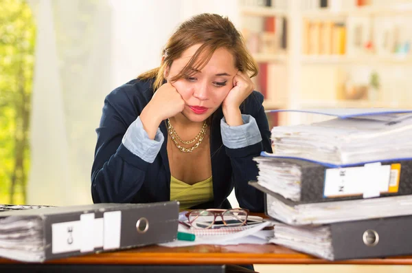 Business woman sitting by desk, paper files spread out, leaning head on hands looking overwhelmed and tired — Stock Photo, Image