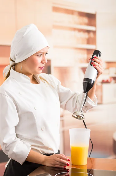 Női szakács kezében elektromos turmixgép és keres szkeptikus, sárga folyadék belsejében magas üveg — Stock Fotó