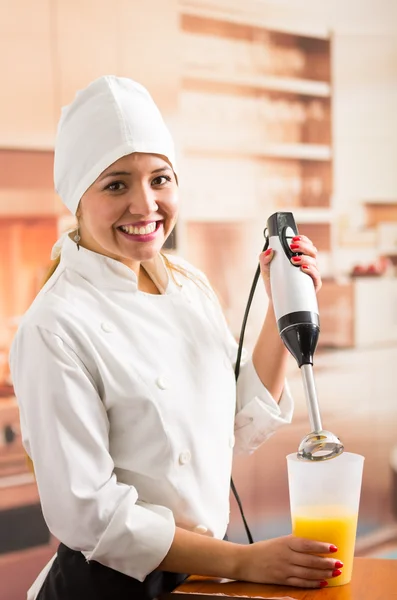 Kvinna kock ler mot kameran med hjälp av handhållen elektrisk mixer med gul vätska inuti högt glas — Stockfoto