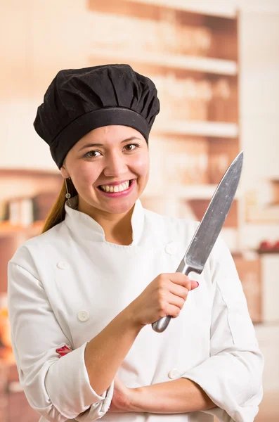 Köchin im Kochkostüm posiert glücklich mit großem metallenen Küchenmesser — Stockfoto