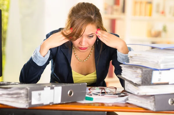 Business woman sitting by desk, paper files spread out, elbows on table and head bent over as expressing great frustration — Stock Photo, Image