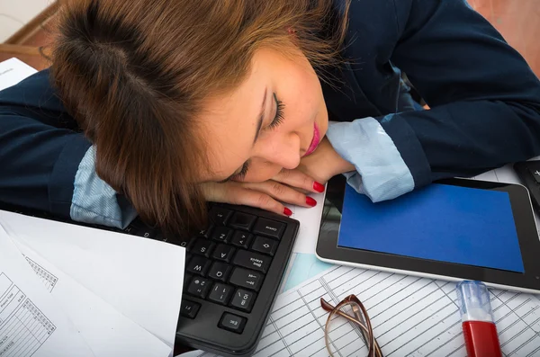 Young woman sitting and lying asleep over office desk with papers, calculator, pens, computer keyboard — Stock Photo, Image