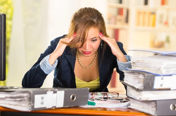 Business woman sitting by desk, paper files spread out, elbows on table and head bent over as expressing great frustration — Stock Photo, Image