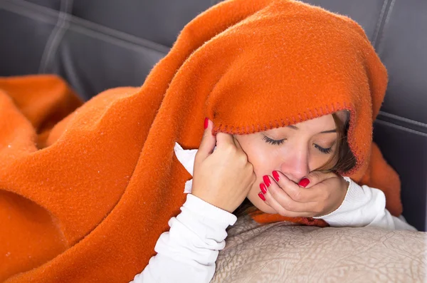 Morena acostada debajo de una manta naranja y cubriéndose la boca con la mano, enferma de gripe — Foto de Stock