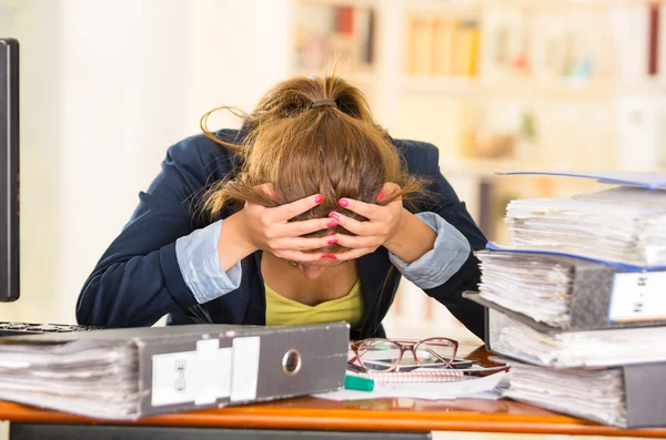 Business woman sitting by desk, paper files spread out, elbows on table and head bent over as expressing great frustration — Stock Photo, Image