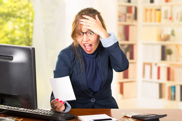 Business woman wearing glasses sitting by computer desk looking at piece of paper with shocked facial expression — Stock Photo, Image