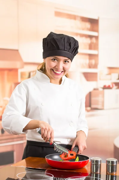 Mujer chef con traje de cocina completo de pie freír verduras en sartén roja y sonriendo a la cámara —  Fotos de Stock