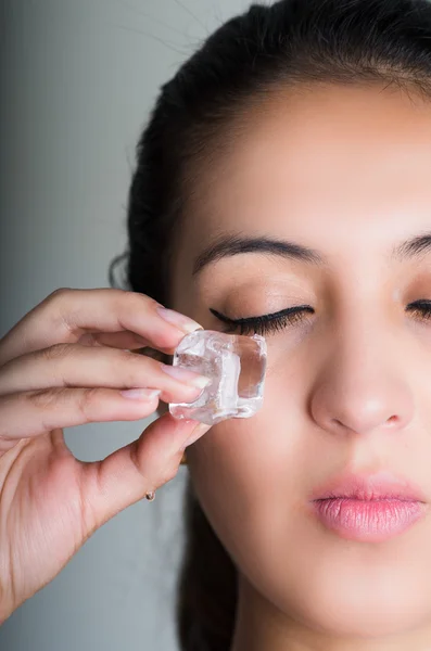 Hermosa modelo morena frotando cubo de hielo contra la mejilla debajo del ojo, fondo gris — Foto de Stock