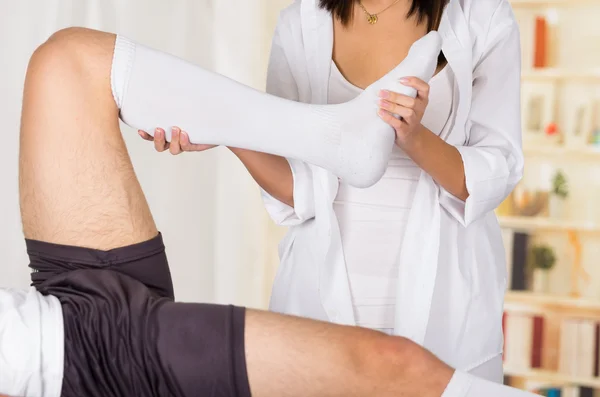 Manos fisioterapeutas femeninas trabajando en pacientes masculinos pierna y tobillo, rodilla doblada, fondo de clínica borrosa —  Fotos de Stock