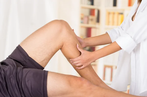 Manos fisioterapeutas femeninas trabajando en las piernas de los pacientes masculinos, sosteniendo y doblando, fondo de la clínica borrosa — Foto de Stock