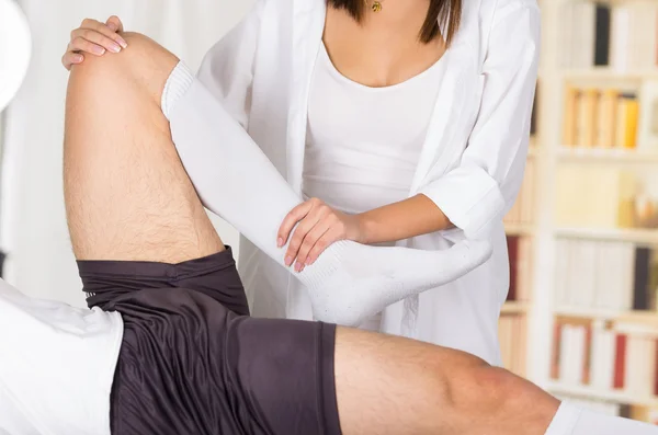 Mulheres fisioterapeuta mãos trabalhando em pacientes do sexo masculino perna e tornozelo, flexão do joelho, fundo clínica embaçada — Fotografia de Stock