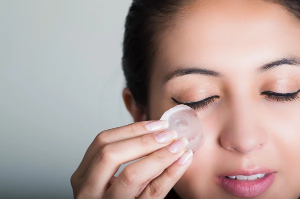 Hermosa modelo morena frotando cubo de hielo contra la mejilla debajo del ojo, fondo gris — Foto de Stock