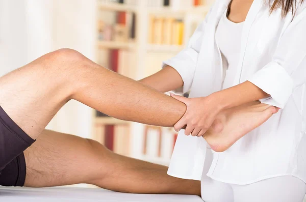 Physiothérapeute féminine mains travaillant sur les patients masculins jambe et cheville, arrière-plan de clinique floue — Photo