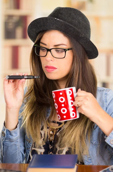 Vrouwelijke brunette hipster Typ dragen bril, hoed en denim jasje zitten door tabel met boek terwijl roken e-sigaret, houden van rode koffiemok — Stockfoto