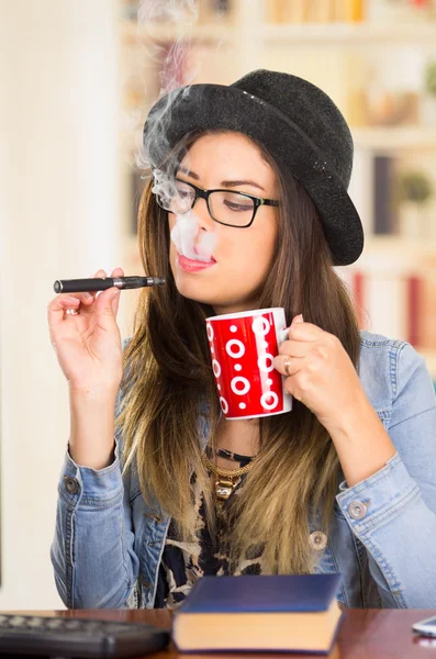 Tipo hipster bruna donna con occhiali, cappello e giacca di denim seduta accanto al tavolo con libro mentre fuma e-sigaretta, con in mano una tazza di caffè rosso — Foto Stock