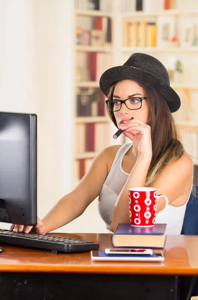 Jeune employée de bureau brune à la hanche portant des lunettes et un chapeau à la mode, assise près du bureau avec un ordinateur tout en fumant une cigarette électronique, tasse à café rouge placée sur des livres — Photo