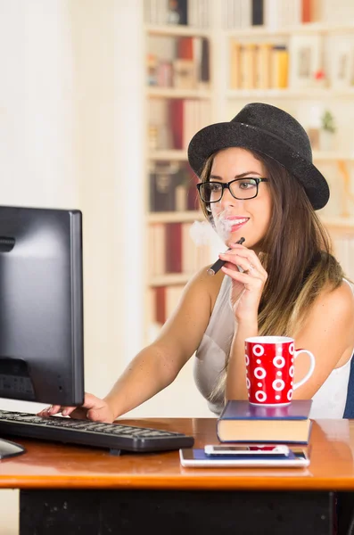 Young hip brunette office worker wearing glasses and fashionable hat, sitting down by desk with computer while smoking e-cigarette, red coffee mug placed on books