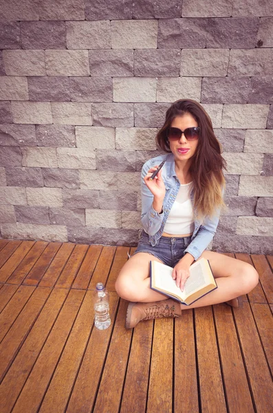 Giovane bruna seduta gambe incrociate su una superficie di legno appoggiata al muro di pietra, fumando e-sigaretta felicemente e leggendo libro — Foto Stock