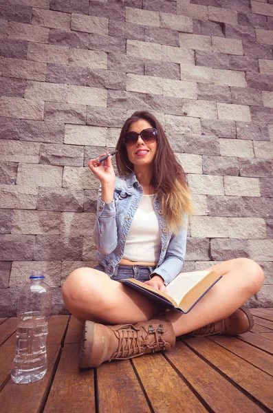 Young hip brunette sitting legs crossed on wooden surface leaning back towards stone wall, smoking e-cigarette happily and reading book — Stock Photo, Image