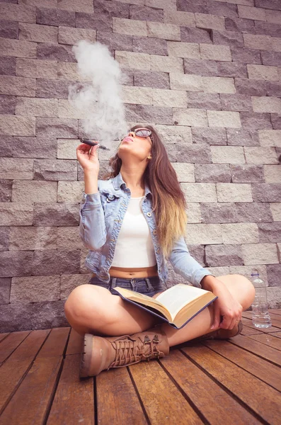 Young hip brunette sitting legs crossed on wooden surface leaning back towards stone wall, smoking e-cigarette happily and reading book — Stock Photo, Image