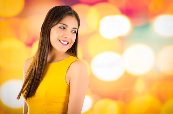 Atractiva morena con vestido amarillo posando naturalmente y sonriendo hermosa a la cámara con fondo colorido borroso —  Fotos de Stock