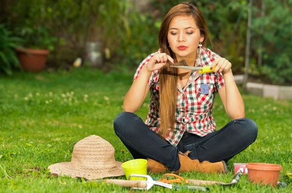穿着牛仔裤和正方形衬衫的年轻妇女在户外工作时，用园艺工具坐在草地上 — 图库照片