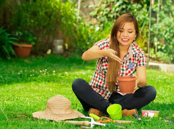 Jeune femme portant un jean et chemise à motif carré assis sur l'herbe tenant pot de fleurs travaillant à l'extérieur — Photo