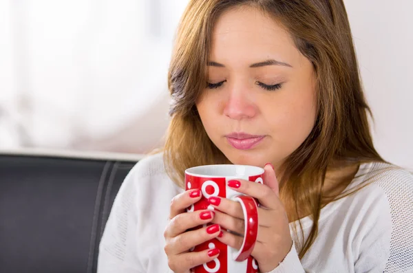 Brunette sick with the flu sitting in sofa holding red cup, tired facial expression — Stock Photo, Image