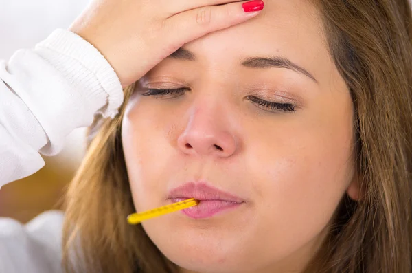 Closeup headshot brunette sick with the flu holding her head, thermometer in mouth — Stock Photo, Image