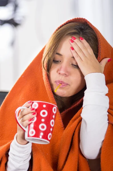 Morena sentada debajo de una manta naranja, sosteniendo la taza roja en la mano, enferma con el concepto de gripe — Foto de Stock