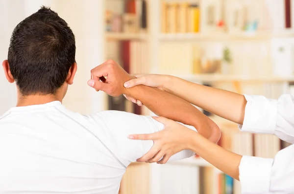 Female physio therapist and man seen from behind, helping patient stretch arm behind head, blurry clinic background