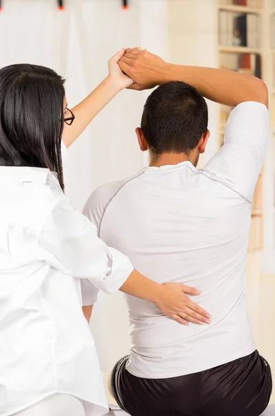 Fisioterapeuta y hombre visto por detrás, ayudando a la paciente a estirar el brazo detrás de la cabeza, fondo de la clínica borrosa —  Fotos de Stock