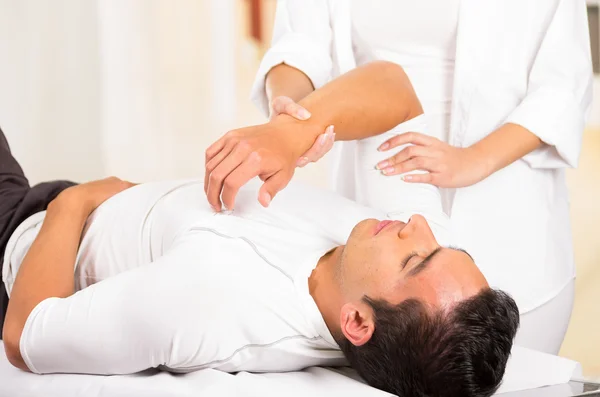 Paciente masculino acostado con manos fisioterapeutas femeninas realizando algunos ejercicios de estiramiento en el brazo del hombre — Foto de Stock