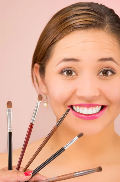 Headshot giovane bella donna ispanica bruna con rossetto rosso che regge la selezione di pennelli per il trucco, sfondo rosa — Foto Stock