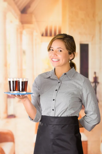 Giovane cameriera bruna indossa uniforme con sorriso amichevole, tenendo in mano vassoio contenente due bicchieri di liquido scuro con un braccio — Foto Stock