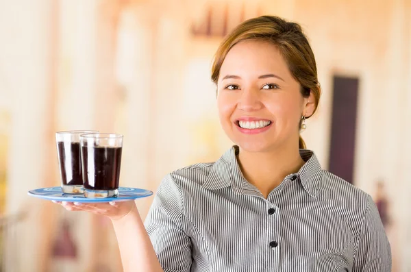 Joven camarera morena vistiendo uniforme con sonrisa amistosa, sosteniendo bandeja que contiene dos vasos de líquido oscuro usando un brazo — Foto de Stock