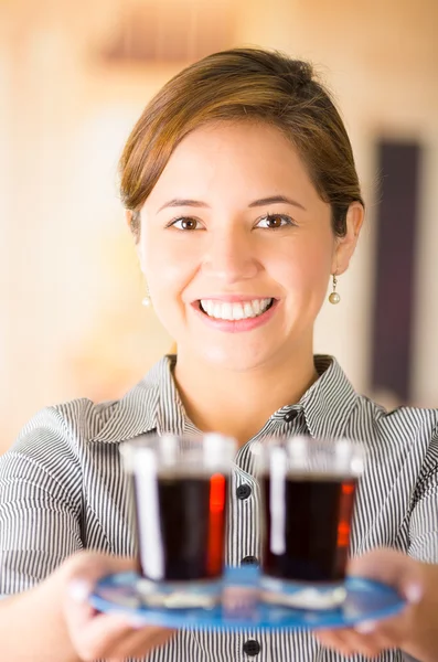 Close-up jonge brunette serveerster uniform dragen met vriendelijke glimlach, het houden van lade met twee glazen donkere vloeistof met behulp van beide handen — Stockfoto