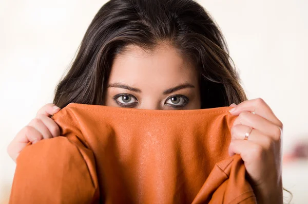 Headshot aantrekkelijke brunette geconfronteerd met camera dekking van de helft van haar gezicht met oranje kleding, witte studio achtergrond — Stockfoto