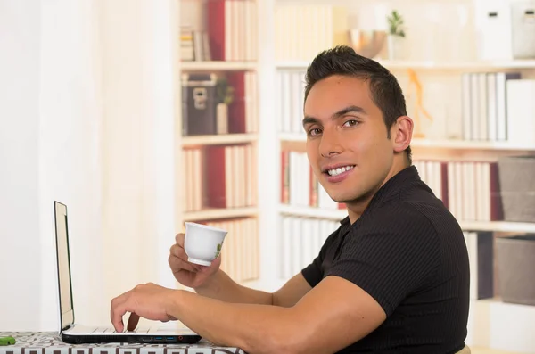 Joven hombre hispano sentado en el escritorio sosteniendo una pequeña taza de café expreso y trabajando en una computadora portátil blanca, ángulo de perfil —  Fotos de Stock