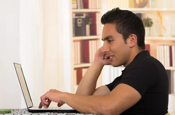 Jeune homme hispanique assis au bureau travaillant sur ordinateur portable blanc, angle de profil — Photo