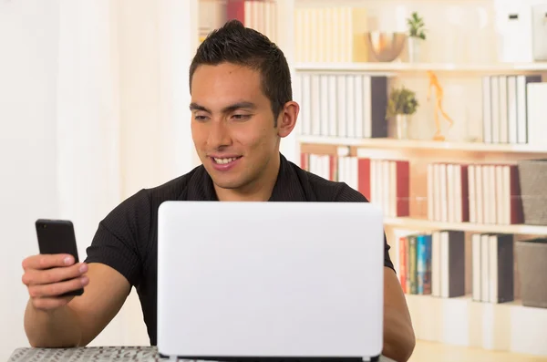 Junger hispanischer Mann sitzt am Schreibtisch und arbeitet an weißem Laptop und schaut aufs Handy — Stockfoto