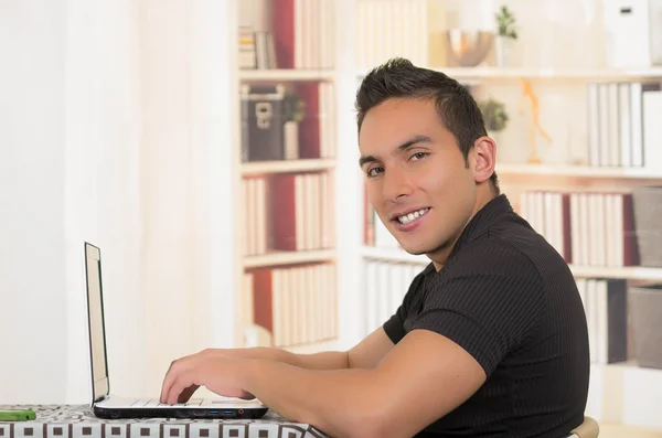 Junger hispanischer Mann sitzt am Schreibtisch und arbeitet an weißem Laptop, Profilwinkel — Stockfoto